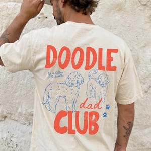 May include: An ivory short-sleeve t-shirt featuring a graphic on the back with the red text "DOODLE DAD CLUB" and "life is better with doodles" in blue script. The design includes two blue line-art dog illustrations and blue paw prints.