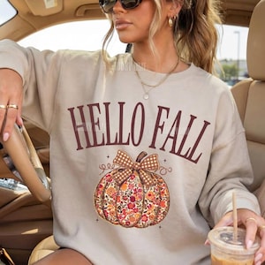 Op de afbeelding: Een zandkleurige sweater met ronde hals en de tekst HELLO FALL in donkerbruine letters boven een afbeelding van een pompoen met een bloemenpatroon en een bruine geruite strik. De sweater heeft lange mouwen en geribbelde boorden.