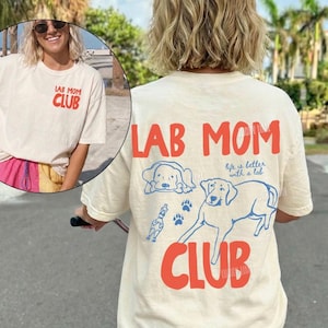 May include: Cream-coloured t-shirt with the text "LAB MOM CLUB" in red. The back of the shirt features a large graphic of a Labrador Retriever and the words "Life is better with a lab". The shirt is a casual style.