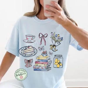 May include: Pale blue t-shirt with a charming illustration of a teacup, cake, bows, butterflies, flowers, books, and a lemon jar. The shirt has the text "Original Design" and "Lucky Lime Co."