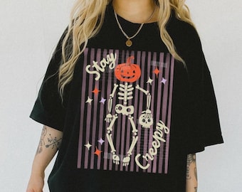 May include: Black t-shirt with a Halloween-themed graphic. The design features a skeleton with a pumpkin head, holding a skull, and the words "Stay Creepy". The background has purple stripes and star accents.