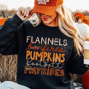 Pode incluir: Uma camisola preta de manga comprida com o texto FLANNELS, bonfires, PUMPKINS, football e HAYRIDES em tons de laranja e creme. A camisola é combinada com um boné de basebol de veludo cotelê laranja com o texto PUMPKIN SPICE.