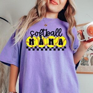 Op de afbeelding: Paars T-shirt met de tekst "Softball Mama" in zwart met gele softball-afbeeldingen en een zwart-wit geruit patroon.
