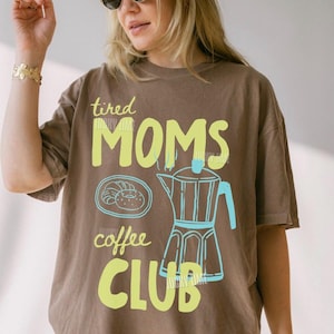 May include: Brown t-shirt with the text "tired MOMS coffee CLUB" in yellow and light blue. The design includes a coffee pot and a pastry. The shirt is a relaxed fit.