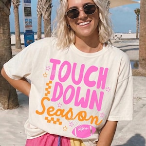 May include: Ivory-colored t-shirt with the words "TOUCH DOWN Season" in pink and orange, with a football graphic. The shirt is paired with pink, yellow, and blue shorts. The design includes stars and a football graphic.
