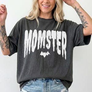 May include: Dark gray t-shirt with the word "MOMSTER" in white, a bat graphic, and the text "Lucky Lime Co". The shirt is a relaxed fit and is paired with denim shorts.