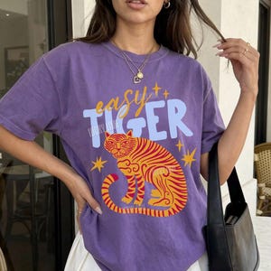 May include: A grape-colored t-shirt with the text "easy tiger" and a tiger graphic in orange and yellow. The shirt is paired with a white pleated skirt and a black shoulder bag. The model is wearing a necklace.