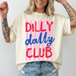 May include: Ivory-colored t-shirt with the text "DILLY DALLY CLUB" in red and blue, set against pink vertical stripes. The shirt is a relaxed fit, paired with denim shorts. The design includes a small logo in the bottom left corner.