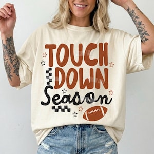 Könnte beinhalten: Elfenbeinfarbenes T-Shirt mit dem Aufdruck "TOUCH DOWN Season" in Braun und Schwarz sowie einer Football-Grafik. Das Shirt hat eine lockere Passform und wird mit Jeansshorts kombiniert. Das Design enthält Sternakzente.