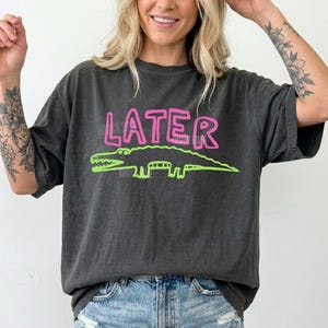May include: Dark grey t-shirt featuring the word "LATER" in pink lettering and a green alligator design. The t-shirt is paired with denim shorts. The design is printed on a relaxed fit t-shirt.