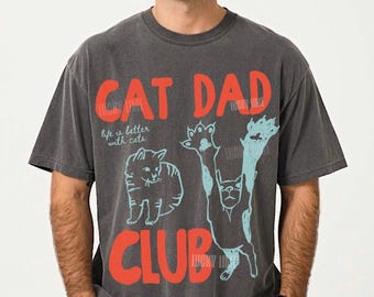 May include: Dark gray t-shirt with the text "CAT DAD CLUB" in red and light blue. The shirt features a cat illustration and the phrase "life is better with cats". The shirt is a relaxed fit.