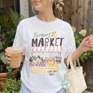May include: White t-shirt with a colorful Farmers Market graphic. The design features a market stall with fruits and vegetables, and the text "Farmers Market" in a stylized font. The shirt also has the text "10AM-5PM DAILY".