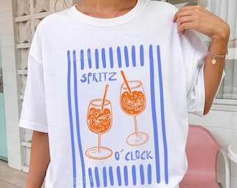 May include: A white short-sleeve t-shirt featuring a graphic print of two orange cocktail glasses with straws, surrounded by vertical blue stripes. The text "SPRITZ O'CLOCK" is printed in blue between the glasses on the front of the garment.