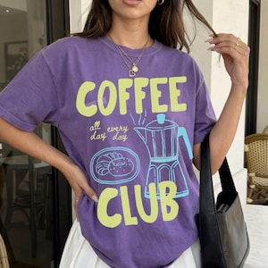 May include: A purple t-shirt with the words "COFFEE CLUB" in a lime green font. The shirt also features a coffee pot and a pastry graphic in light blue. The text "all every day day" is above the coffee pot graphic. The shirt is paired with a white pleated skirt.