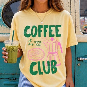Peut inclure: Un t-shirt jaune p&acirc;le &agrave; manches courtes avec le texte vert COFFEE CLUB et un contour rose d'une cafeti&egrave;re italienne avec une p&acirc;tisserie. Le t-shirt porte le texte all every day en petites lettres vertes. Un gobelet en plastique de th&eacute; glac&eacute; vert.