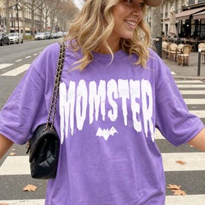 May include: A violet t-shirt with the word "MOMSTER" in large white letters and a bat graphic. The shirt is worn with a black quilted shoulder bag and a beige baseball cap. The word "VIOLET" is printed at the bottom left.