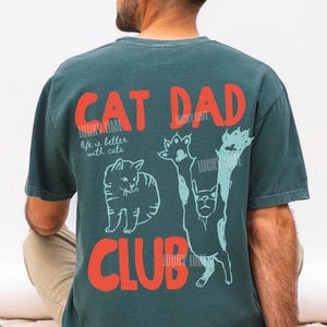 Peut inclure: T-shirt bleu sarcelle avec l'inscription "CAT DAD CLUB" en lettres rouges. Le t-shirt pr&eacute;sente un motif de chat et la phrase "life is better with cats" en blanc. C'est un design Blue Spruce.