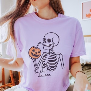 Op de afbeelding: Lila T-shirt met een zwart-wit skelet dat uit een oranje pompoen drinkt. De tekst "'tis the Season" staat eronder. Een casual shirt, perfect voor de herfst of Halloween.