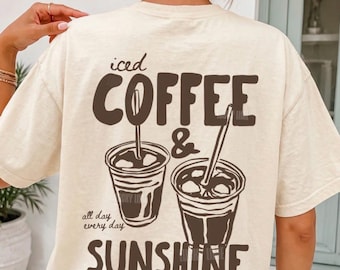 Puede incluir: Una camiseta color marfil con un gráfico marrón oscuro en la parte posterior. El diseño muestra dos vasos de café helado con pajitas y el texto "iced COFFEE & SUNSHINE" junto a la frase "all day every day" en una tipografía casual y negrita.