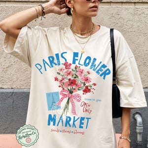 Peut inclure: T-shirt crème oversize avec un motif de marché aux fleurs. Le motif comprend le texte "Paris Flower Market" en bleu, un bouquet de fleurs roses et le texte "Open Daily". Le t-shirt est porté avec un short à carreaux rouges et blancs.