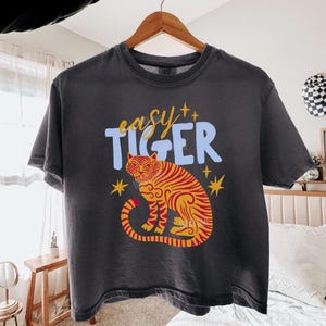 Op de afbeelding: Donkergrijs t-shirt met de tekst "easy tiger" in lichtblauw en geel. Het shirt heeft een gedetailleerde illustratie van een tijger in oranje en geel met steraccenten. Het shirt hangt aan een houten hanger.