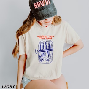 以下が含まれることがあります： クリーム色のTシャツに、青と赤のグラフィックで、サージン缶と「We're in this together」というテキストが描かれています。