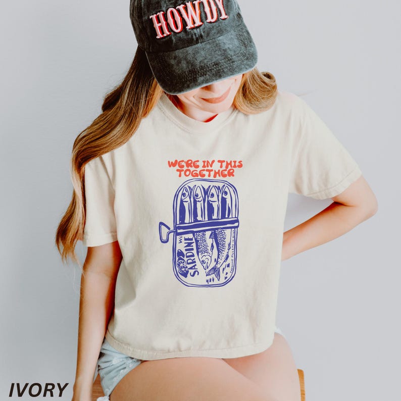 以下が含まれることがあります： クリーム色のTシャツに、青と赤のグラフィックで、サージン缶と「We're in this together」というテキストが描かれています。