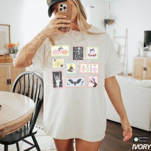 May include: Ivory-colored t-shirt featuring a collection of Halloween-themed postage stamp designs. The stamps showcase pumpkins, skeletons, spiders, witch hats, potion bottles, and bats. The shirt has a relaxed fit and short sleeves.