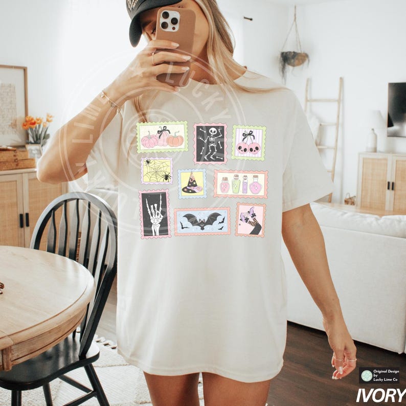 May include: Ivory-colored t-shirt featuring a collection of Halloween-themed postage stamp designs. The stamps showcase pumpkins, skeletons, spiders, witch hats, potion bottles, and bats. The shirt has a relaxed fit and short sleeves.