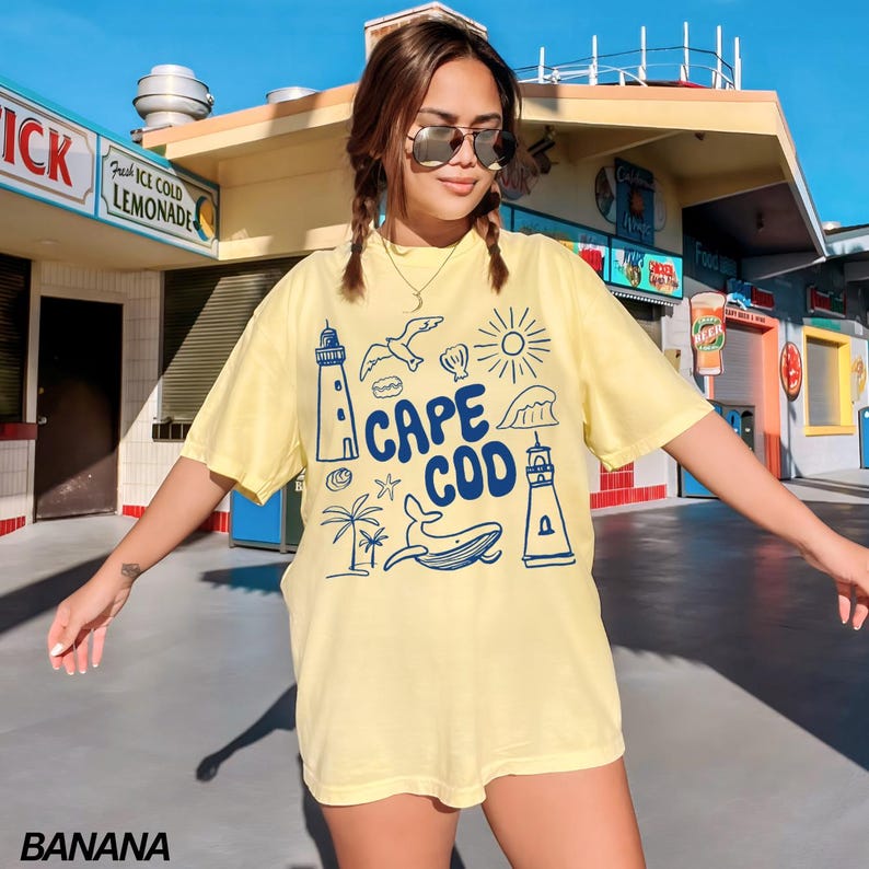 May include: A yellow short-sleeved t-shirt with a blue graphic print of a lighthouse, a whale, a sun, a wave, and the text "Cape Cod".