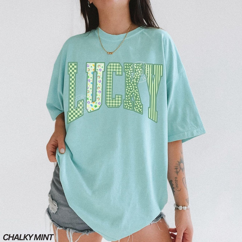 May include: A light blue t-shirt with the word "LUCKY" in green, checkered, and striped patterns. The shirt is a relaxed fit and has short sleeves. The text "CHALKY MINT" is at the bottom.