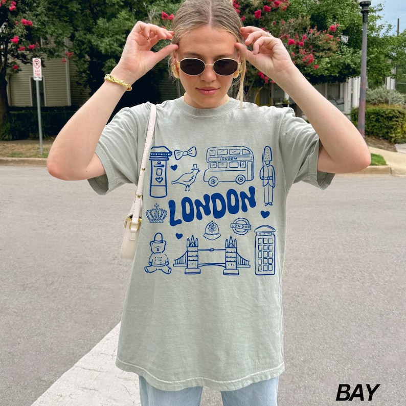 May include: A light green t-shirt with a blue graphic print of London landmarks, including a double-decker bus, a red telephone booth, a crown, and the words "LONDON".