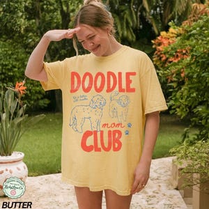 Puede incluir: Camiseta amarilla con el texto "DOODLE mom CLUB" en rojo, con ilustraciones de perros doodle. La camiseta tiene un corte holgado y mangas cortas. La camiseta est&aacute; hecha de un material suave.