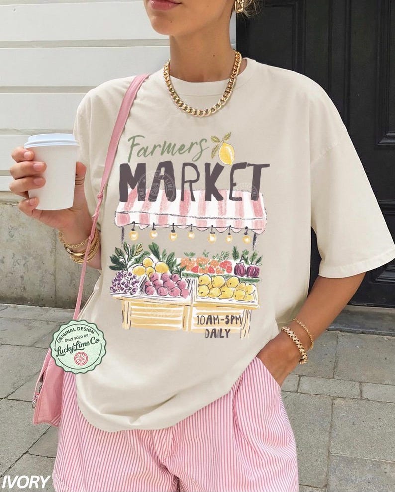 May include: Ivory-colored t-shirt with a Farmers Market graphic. The design features a market stall with fruits and vegetables, the text "Farmers Market", and the hours "10AM-5PM Daily". The shirt is paired with pink striped shorts.