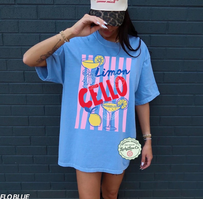 May include: A light blue t-shirt with the words "Limon Cello" in a colorful design. The shirt features a cocktail glass graphic with a lemon slice, set against pink and white vertical stripes. The shirt is a Flo Blue color.