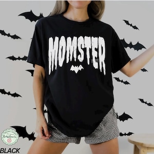May include: Black t-shirt with the word "MOMSTER" in white, featuring a bat graphic. The shirt is part of a Halloween-themed collection. The text "Lucky Lime Co" is also visible. The model is wearing black and white checkered shorts.
