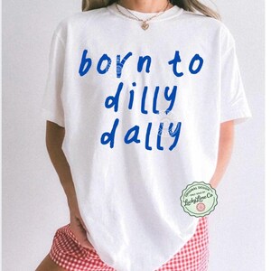 May include: White t-shirt with the blue text "born to dilly dally". The shirt has a round neck and short sleeves. The model is wearing red and white checkered shorts. A small green and white circular logo is on the right side of the shirt.