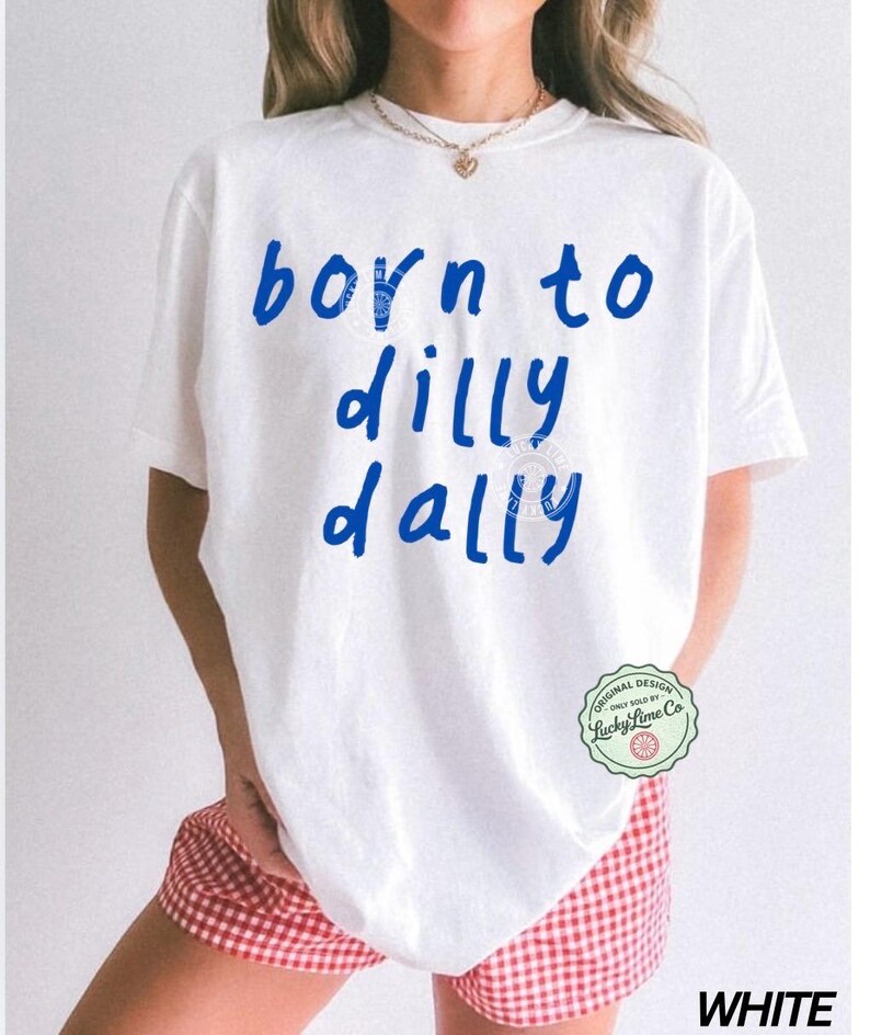 May include: White t-shirt with the blue text "born to dilly dally". The shirt has a round neck and short sleeves. The model is wearing red and white checkered shorts. A small green and white circular logo is on the right side of the shirt.