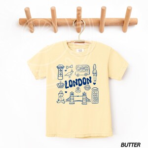 May include: A yellow t-shirt featuring a blue graphic of London landmarks, including a double-decker bus, Big Ben, a red telephone box, and Tower Bridge. The text "LONDON" is printed in blue on the shirt.