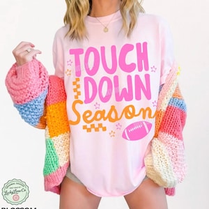 May include: A light pink t-shirt with the words "TOUCH DOWN Season" in pink and orange, along with a pink football graphic. The shirt is paired with a colorful, patchwork cardigan in shades of pink, blue, orange, and yellow.