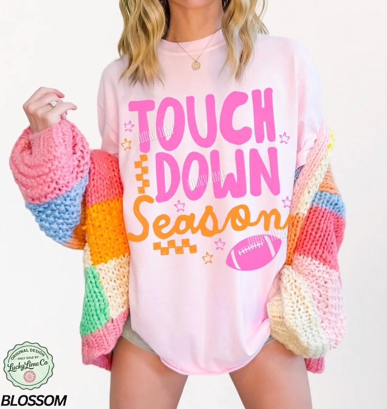 May include: A light pink t-shirt with the words "TOUCH DOWN Season" in pink and orange, along with a pink football graphic. The shirt is paired with a colorful, patchwork cardigan in shades of pink, blue, orange, and yellow.