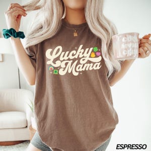 Peut inclure: Un t-shirt marron avec le texte "Lucky Mama" dans une police r&eacute;tro. Le texte est de couleur cr&egrave;me et est entour&eacute; de petits tr&egrave;fles color&eacute;s et d'un arc-en-ciel.