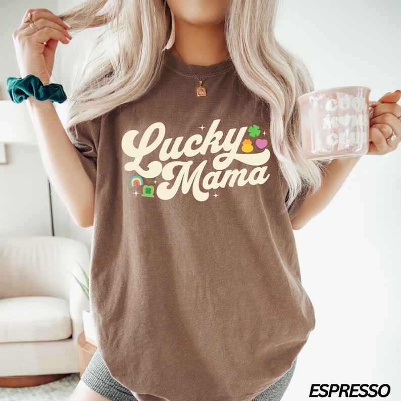 Peut inclure: Un t-shirt marron avec le texte "Lucky Mama" dans une police r&eacute;tro. Le texte est de couleur cr&egrave;me et est entour&eacute; de petits tr&egrave;fles color&eacute;s et d'un arc-en-ciel.