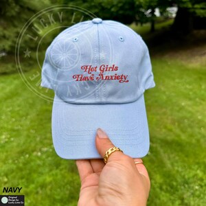 May include: Light blue baseball cap with the text "Hot Girls Have Anxiety" in red script. The cap is made of a soft material and has a curved brim. The cap is navy blue.