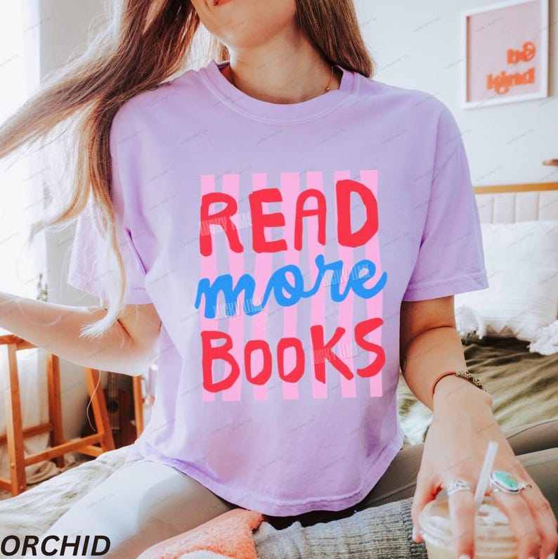 K&ouml;nnte beinhalten: Ein hellviolettes Kurzarm-T-Shirt mit einem Grafikdruck aus vertikalen rosa Streifen. Der Text READ ist in Rot, more in Blau und BOOKS in Rot gedruckt. Das T-Shirt hat einen Rundhalsausschnitt und eine lockere Passform.