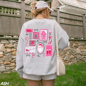 May include: Ash-colored sweatshirt with a festive design. The back features a collage of pink and gray Christmas-themed illustrations, including Santa Claus, a house, and candy canes. The word "HO HO HO" is also visible.