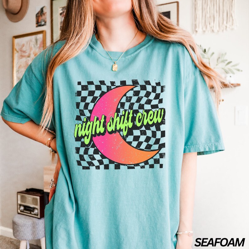 May include: A turquoise blue t-shirt with a graphic of a pink and orange crescent moon on a black and white checkered background. The text "night shift crew" is written in green on the moon.