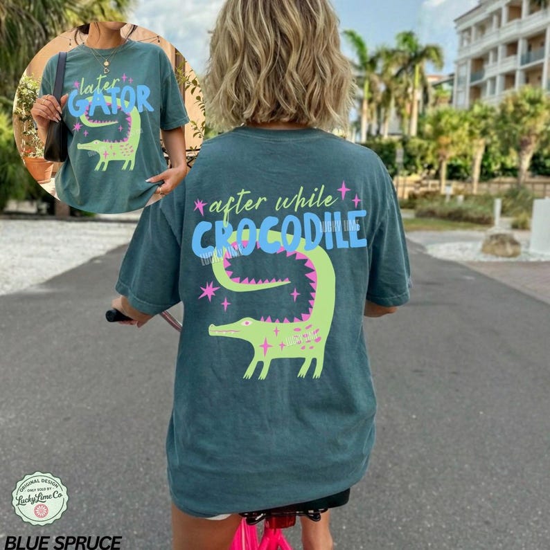 May include: Blue spruce t-shirt with a green alligator graphic and the text "later GATOR" on the front. The back of the shirt features the text "after while CROCODILE" and a larger alligator design. The shirt is a comfortable, casual style.