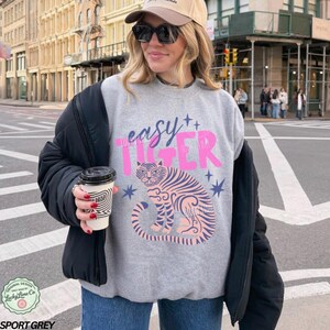May include: Grey sweatshirt with the words "easy TIGER" in pink and blue. It features a pink tiger illustration with blue stripes and stars. The design is from Lucky Lime Co.