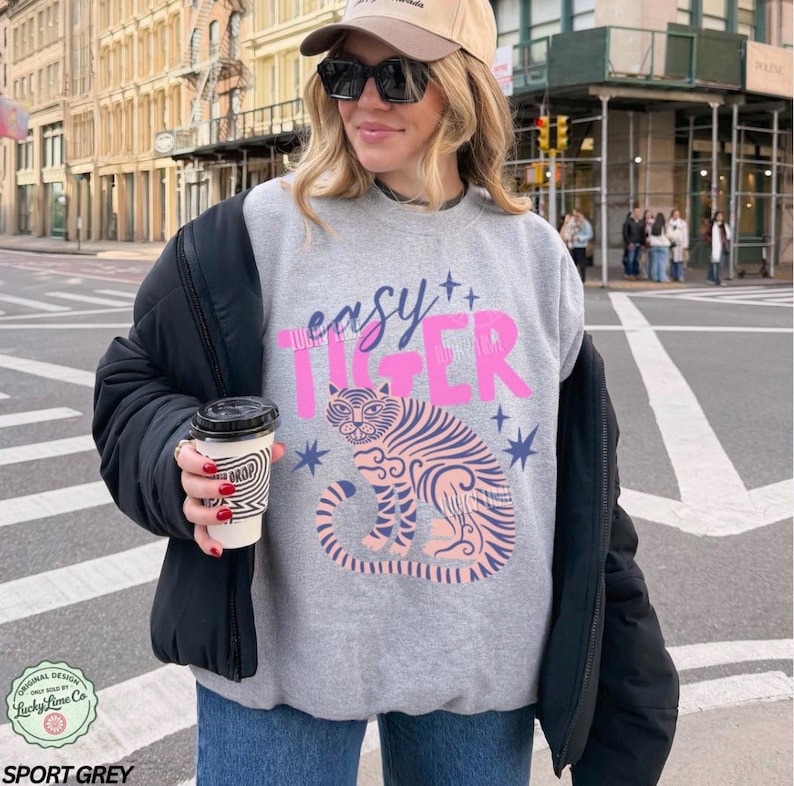 May include: Grey sweatshirt with the words "easy TIGER" in pink and blue. It features a pink tiger illustration with blue stripes and stars. The design is from Lucky Lime Co.